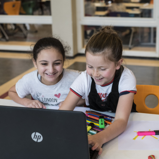 Twee meisjes van de basisschool zitten samen aan een tafel en werken lachend op een laptop. Op tafel liggen stiften en een creatieve opdracht.