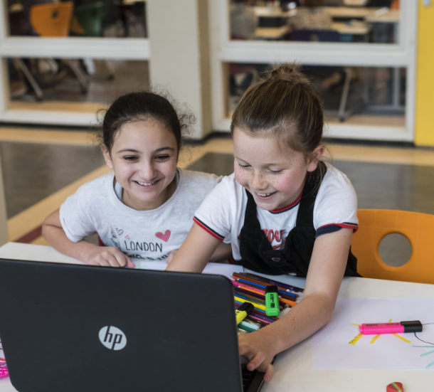 Twee meisjes van de basisschool zitten samen aan een tafel en werken lachend op een laptop. Op tafel liggen stiften en een creatieve opdracht.