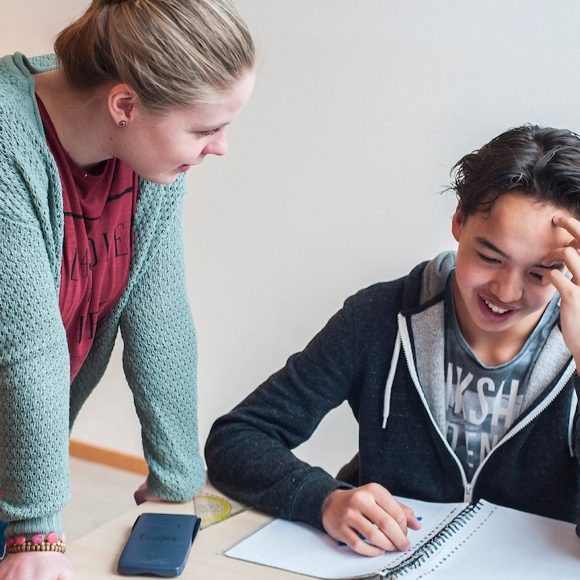 Een jongere zit aan tafel en lacht terwijl hij aan zijn huiswerk werkt. Een begeleider of docent buigt zich vriendelijk naar hem toe om te helpen of mee te denken.
