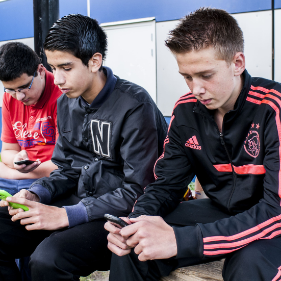 Drie jongens zitten buiten op een houten bank en kijken geconcentreerd op hun telefoon. Ze lijken verdiept in hun scherm, elk met een eigen toestel.
