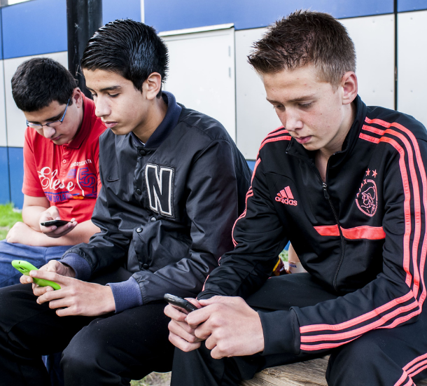 Drie jongens zitten buiten op een houten bank en kijken geconcentreerd op hun telefoon. Ze lijken verdiept in hun scherm, elk met een eigen toestel.