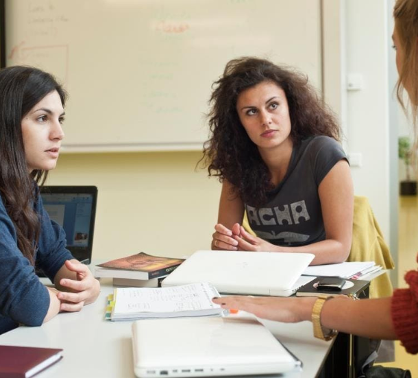 Drie jonge vrouwen zitten samen aan tafel met laptops, boeken en notities. Ze zijn geconcentreerd in gesprek, mogelijk over een studieopdracht of project.